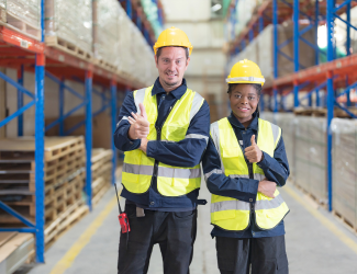 Two warehouse workers standing side by side in a row of stacked pallets wearing yellow safety vests